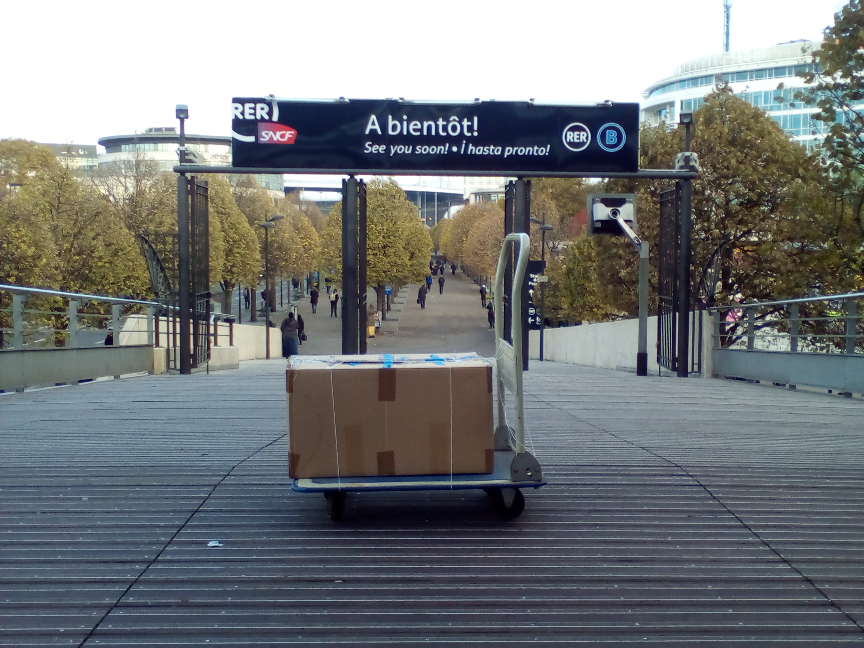 Une boîte en carton posée sur un diable devant un panneau qui déclare : « RER SNCF • À bientôt! • See you soon! • ¡ hasta pronto<span style=